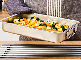 A person wearing an oven mitt holds a stainless steel roasting pan filled with assorted roasted vegetables over a metal pot stand on a wooden table.