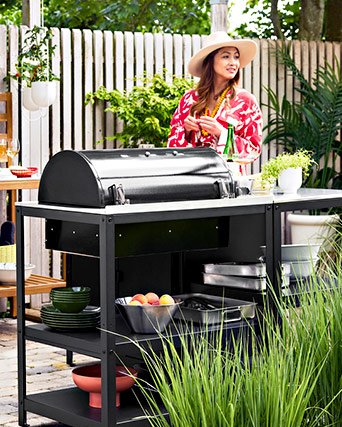A woman stands by a black outdoor charcoal grill on a metal trolley with shelves holding green plates, steel bowls, and a roasting pan.