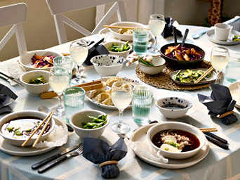 A dining table set with a checkered cloth, featuring plates of dumplings, edamame, soup bowls, and glasses of juice with white chairs.