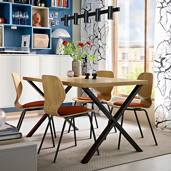 Modern dining area featuring a wooden table with black legs, four chairs with orange cushions, a black linear pendant lamp, and blue wall shelves.