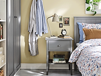 Un rincón de dormitorio con una mesita de noche GULLABERG junto a la cama, mostrada con una lámpara, libros y pequeños objetos en su superficie.