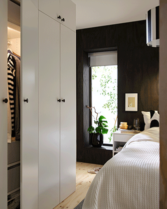 Combinación de armario LASTARE en color blanco con la puerta parcialmente abierta mostrando ropa colgada, situado junto a una cama en un dormitorio moderno.