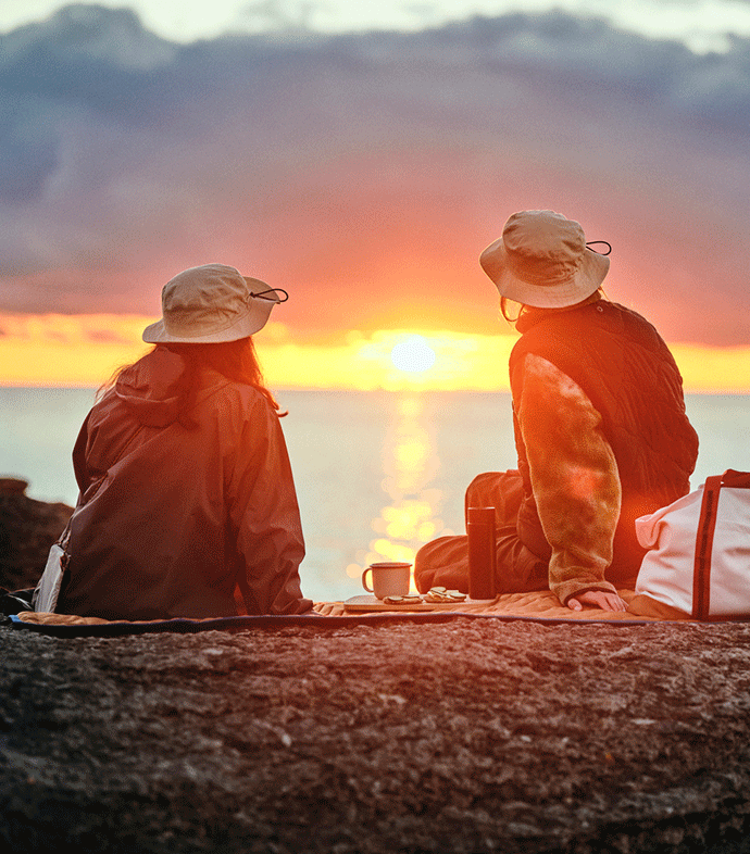 Dos personas que llevan sombreros SOLUPPGÅNG están sentadas sobre una manta de picnic SOLUPPGÅNG mientras ven el atardecer junto al mar.