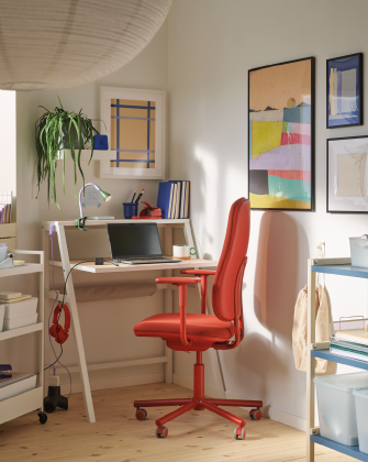 A workspace corner featuring a small white desk, a red office chair, and colorful art prints on the wall.