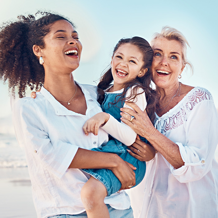 Three generations of women laughing together on a sunny beach. A young girl is held in their arms with pure happiness.