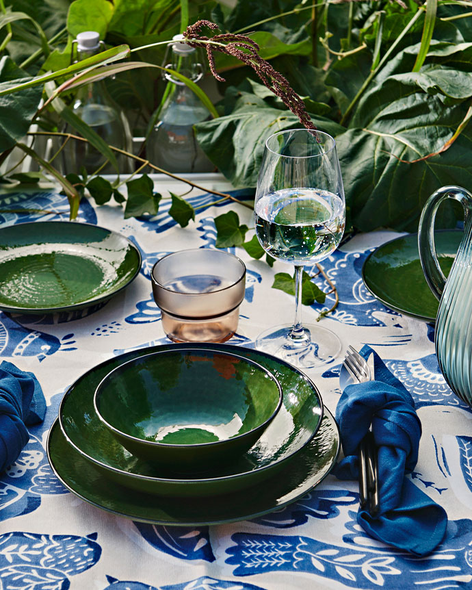 Mesa de exterior con platos y cuencos verdes sobre un mantel blanco con pájaros azules, rodeada de plantas y copas de agua.