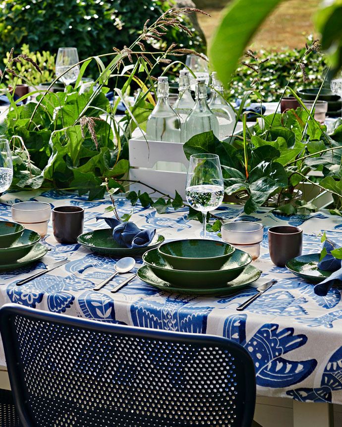 Mesa de jardín decorada con vajilla verde y mantel de pájaros azules. Incluye botellas de vidrio en una caja blanca y sillas.