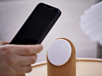A hand holds a black smartphone in front of a white circular wireless charger mounted on a natural cork base.