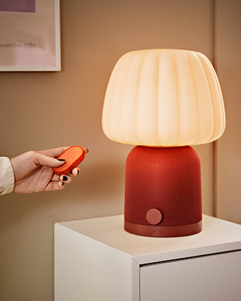 A hand uses an orange remote to control a table lamp featuring a pleated shade and a red speaker base on a white cabinet.