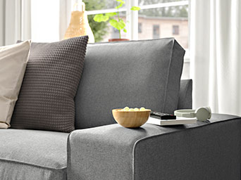 Close-up of a grey sofa armrest with a waffle-texture pillow, a small wooden bowl of snacks, a remote, and headphones on the side.
