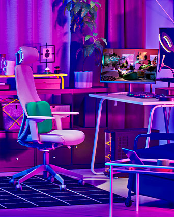 Zona de juegos con iluminación púrpura y azul, una silla ergonómica blanca con cojín verde, un escritorio con monitor y estanterías al fondo.
