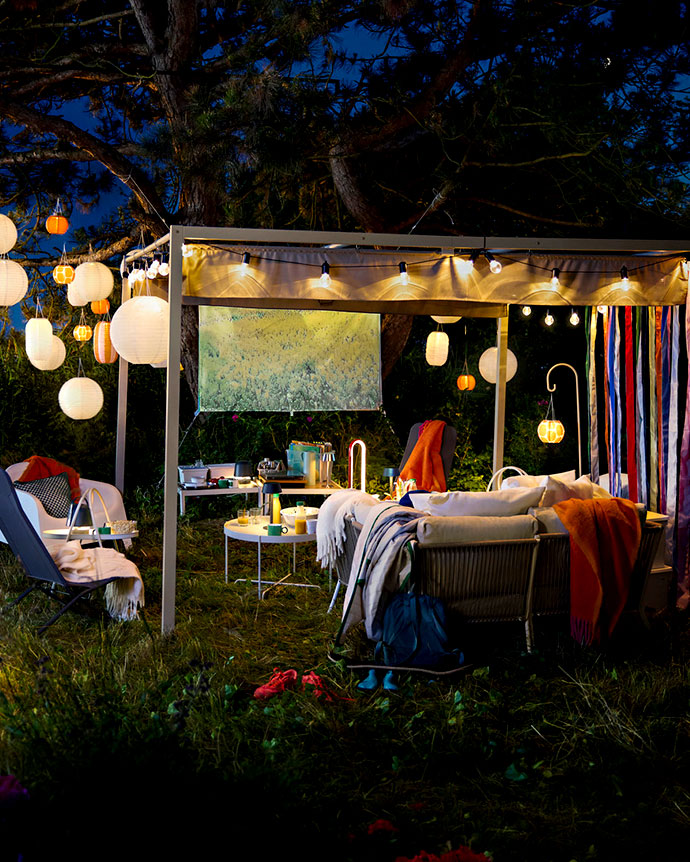Outdoor gazebo at night with paper lanterns, string lights, and cozy sofas with blankets on a grassy garden area.