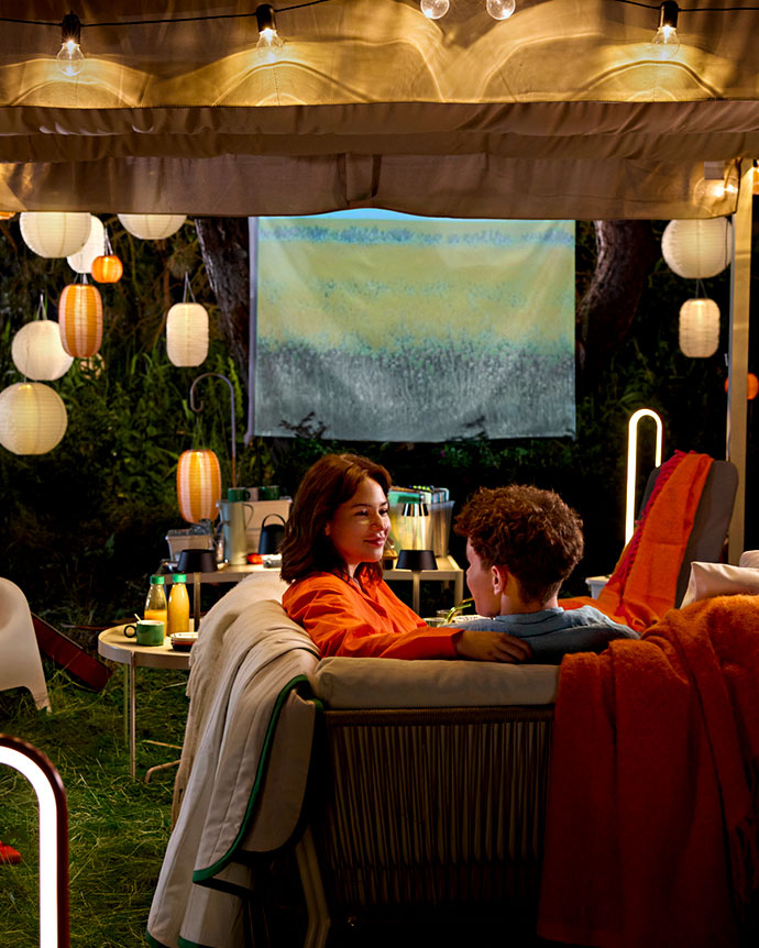 Couple relaxing on a sofa under a lit gazebo, enjoying an outdoor movie projection surrounded by hanging paper lanterns in the garden.