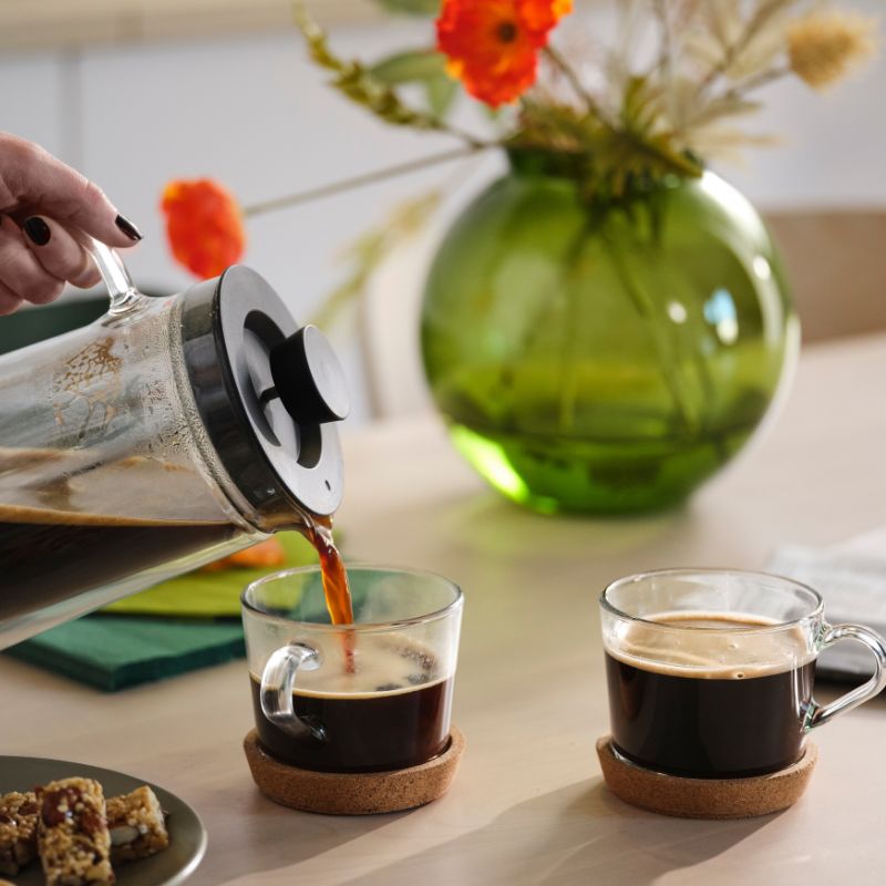 Mesa de café minimalista con taza y posavasos IKEA 365+, cafetera UPPHETTA, plato MOSSMAL y barrita de cereal.