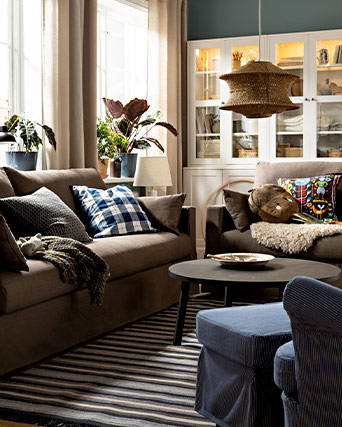 Cozy living room featuring brown sofas, decorative pillows, a round coffee table, and an illuminated white display cabinet. Warm, tidy atmosphere.