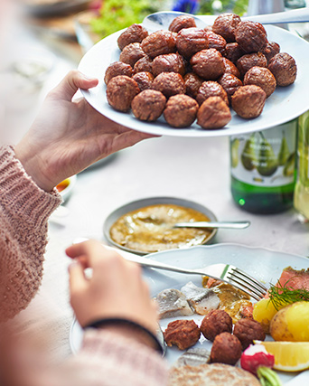 Una mano sosteniendo albóndigas HUVUDROLL en un plato, y otro plato con SILL DILL preparado, junto a un juego de cubiertos DRAGON.