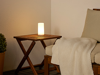 La lámpara decorativa SOLVINDEN sobre una mesa plegable NÄMMARÖ, junto a un sofá modular NÄMMARÖ con una manta HUMLEMOTT encima.