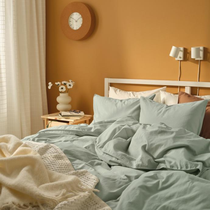 A cozy bedroom with sage green bedding, a cream knit blanket, and a warm mustard-yellow wall with a round wooden clock.