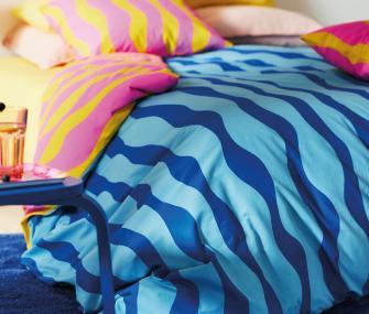 A close-up of a bed with vibrant bedding, featuring bold blue and light blue wavy patterns and pink and yellow pillows.