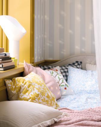A cozy bed corner with multiple decorative pillows in pink, blue, and yellow floral prints against a striped wall and yellow pillar.