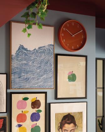 A gallery wall with framed prints of blue waves and colorful apples on a blue wall next to an orange clock.