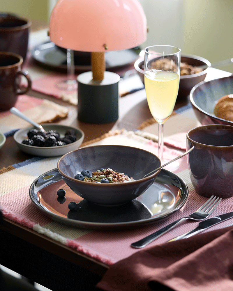 Un cuenco con yogur, muesli y arándanos está colocado junto a una taza GLADELIG de color lila y una copa de champán.