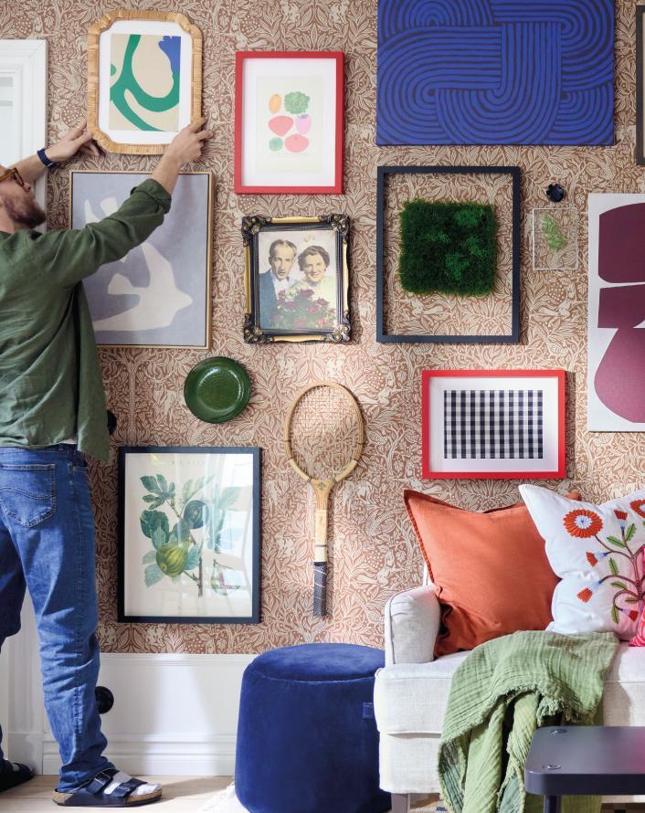 Person arranging framed artwork and decorations on a patterned wallpaper wall in a cozy and colorful living room.