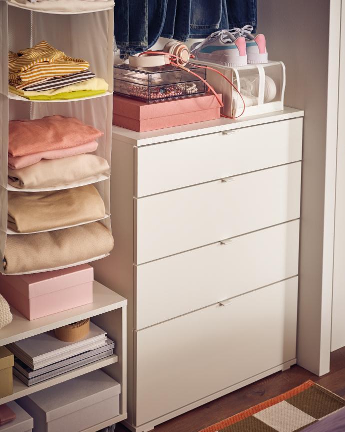 A narrow white chest of drawers inside a modern closet, surrounded by neatly folded clothes, storage boxes, headphones, and sneakers arranged on shelves.
