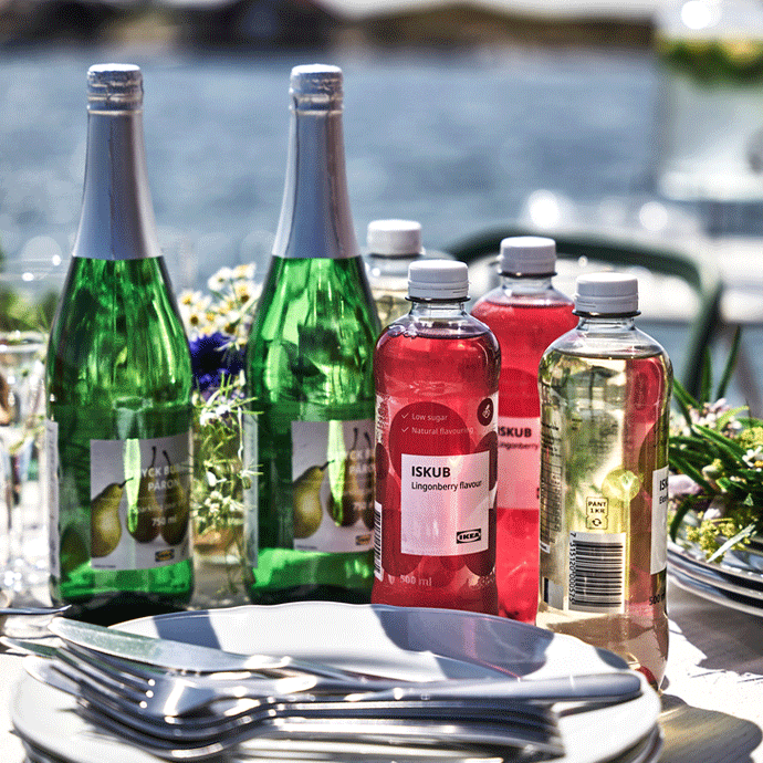 Mesa exterior con botellas verdes de bebida de pera DRYCK BUBBEL PÄRON, botellas de agua ISKUB rojas y amarillas, platos blancos apilados y cubiertos.