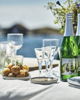Botellas de vidrio verde de la bebida de pera DRYCK BUBBEL PÄRON junto a copas elegantes y un cuenco con patatas cocidas sobre una mesa blanca soleada.