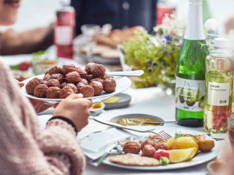 Manos sirviendo un plato lleno de albóndigas suecas sobre una mesa exterior equipada con sidra DRYCK BUBBEL PÄRON, bebidas ISKUB, platos y flores frescas.