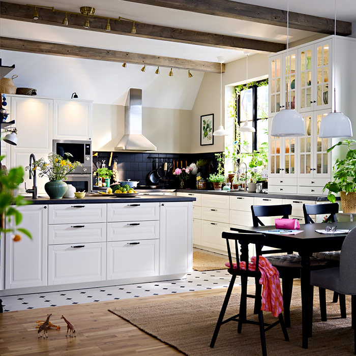 Spacious, bright kitchen featuring white cabinets, a central island, wooden ceiling beams, a black dining table, and plants by the window.
