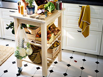 Light wood kitchen kitchen island cart with three tiers, filled with fresh vegetables in crates and a mesh bag, on a tiled floor.
