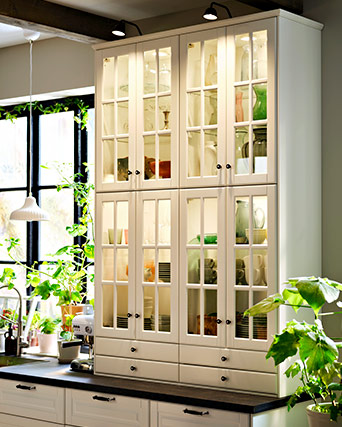Tall white kitchen cabinet with illuminated glass doors showing organized tableware, placed on a dark countertop near a window.