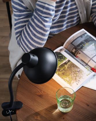 Overhead view of a black clip-on lamp with a round shade illuminating an open book on a wooden table, next to a glass of water and a person wearing a striped sweater.
