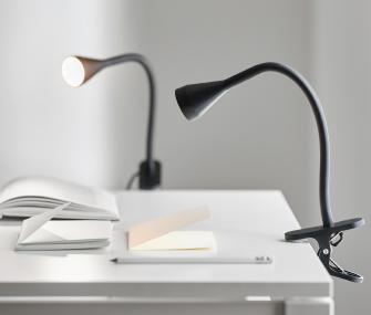 Two minimalist black clip-on reading lights attached to a white desk with notebooks and a pen. The lamp in the background is turned on.