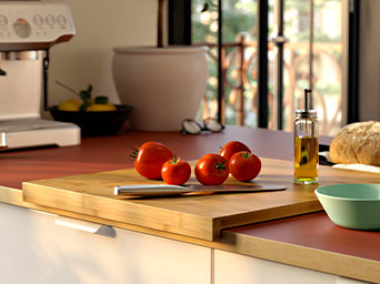 Primer plano de una tabla de cortar de bambú con tomates frescos y un cuchillo sobre una encimera rojiza, con una cafetera al fondo.