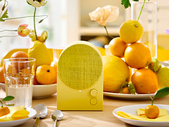 Altavoz amarillo en una mesa decorada con cítricos y flores en tonos cálidos, junto a vasos de agua y cubiertos metálicos.