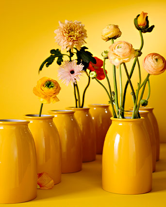 Varios jarrones de cerámica de color amarillo brillante con flores variadas sobre un fondo del mismo tono.