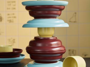 Un juego de servicio de 3 piezas SPJUTLILJA con cuencos morados oscuros, tazas amarillas y platos azul claro, apilados en una mesa oscura.