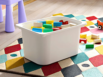 A white plastic storage box filled with colorful wooden toy blocks, on a kids' rug with multicolored diamonds.