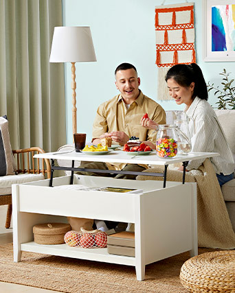 A smiling couple in a living room by a white lift-top coffee table with food, fruit, and a candy jar, set on a natural jute rug.