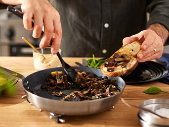 Close-up of a person cooking mushrooms in a frying pan and serving them with a spatula onto a slice of toasted bread.