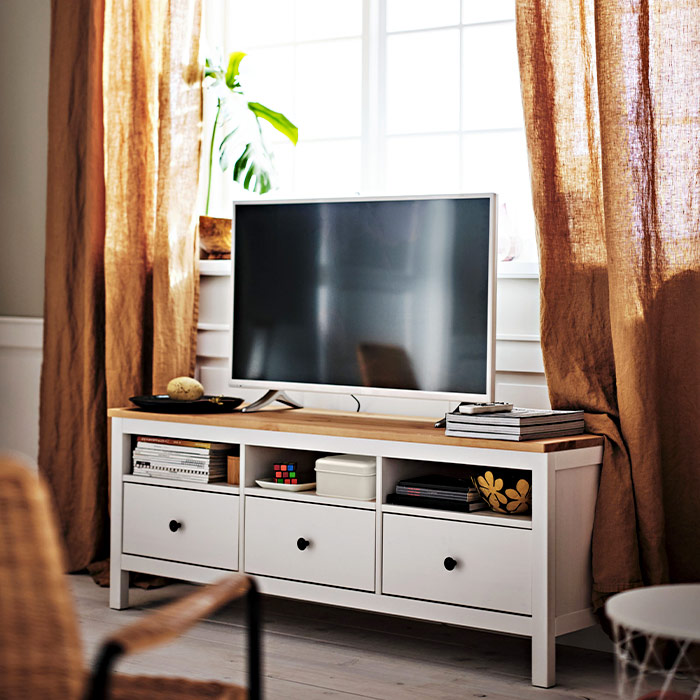 A white TV bench with a wooden top, drawers, and shelves, placed in front of a window with brown linen curtains.