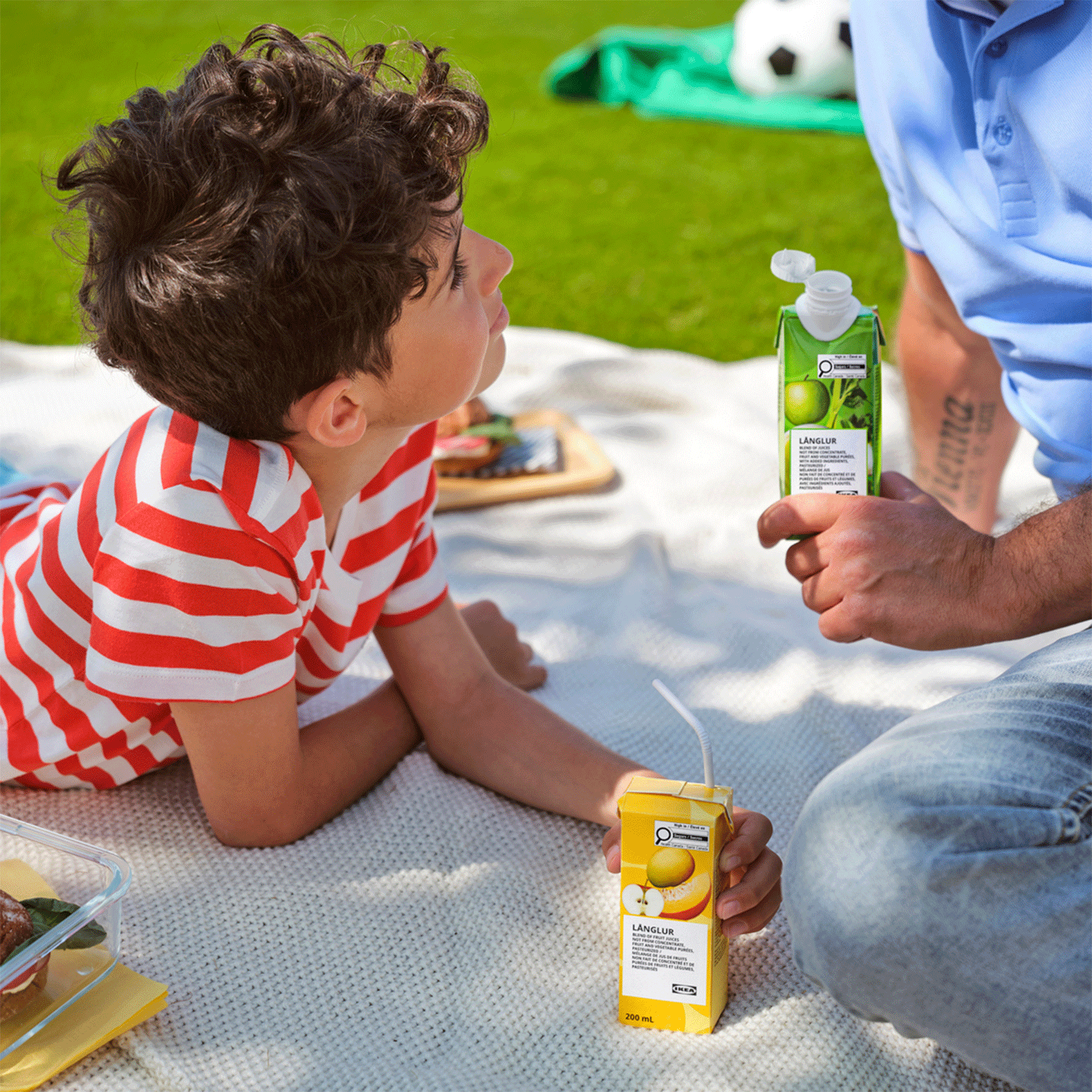 A child in a red and white striped shirt lies on a picnic blanket next to an adult holding an open green LÄNGLUR juice carton, with a yellow juice box on the blanket.