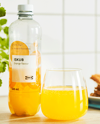 Clear plastic bottle of ISKUB orange flavour drink next to a stemless glass filled with the same vibrant orange, sparkling beverage on a light wooden table.