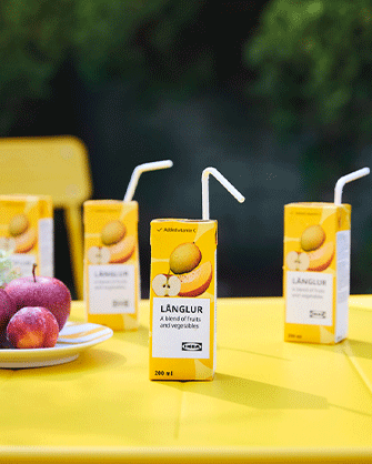 Several yellow 200 ml LÄNGLUR juice cartons with white straws arranged on a bright yellow table next to a white plate containing fresh red apples.