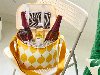 Yellow and white lemon-patterned cooler bag sitting on a white chair, packed with ice cubes, two drink bottles, and two dark glass bottles with red foil wrappers.