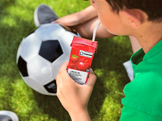 Close-up of a person holding a red LÄNGLUR juice box with a white straw, sitting on green grass next to a classic black and white soccer ball.