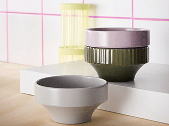 Tres cuencos de cerámica moderna en tonos gris, verde oliva y rosa pálido, expuestos sobre una plataforma blanca con un fondo de azulejos rosas.